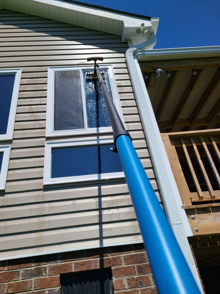 A person cleans a window on a house with a long, blue pole. The house has tan siding, white trim, and a wooden deck.