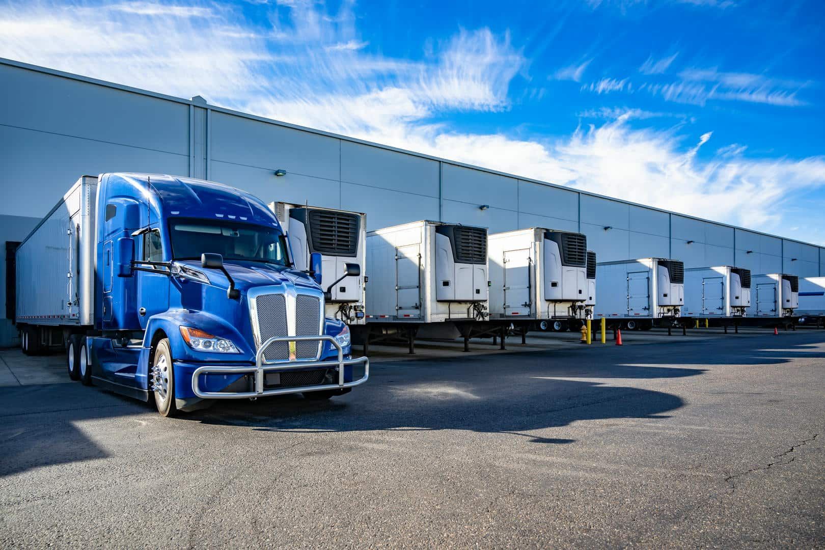 Blue semi-truck parked at loading dock with several refrigerated trailers against a white warehouse, under a blue sky.