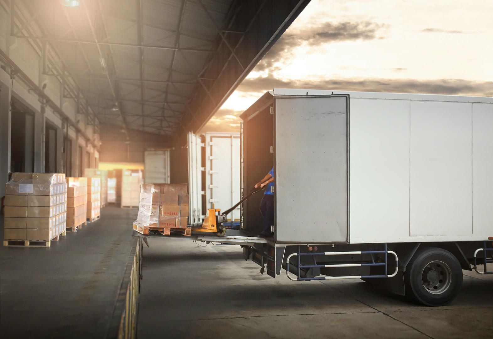 Truck unloading packages at a warehouse loading dock; boxes on pallets, worker, sunlight.