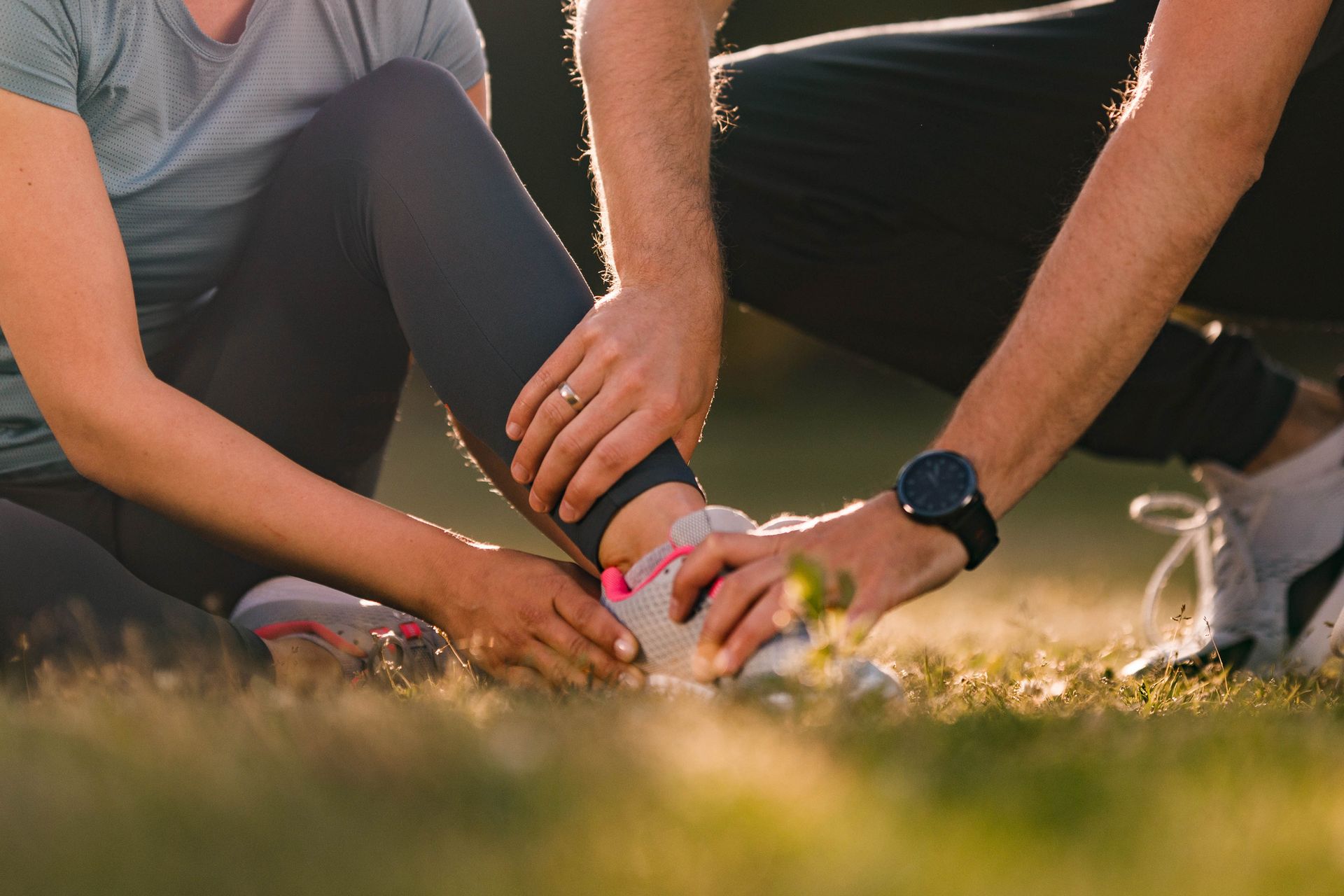 A man is helping a woman tie her shoes while sitting on the grass.