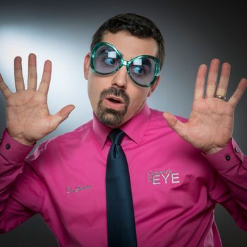 Man wearing large blue sunglasses, pink shirt, and dark blue tie, hands up in surprise.