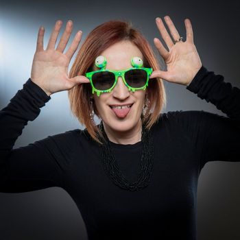 Woman with red hair wearing silly green sunglasses and making a goofy face. She is in front of a gray background.