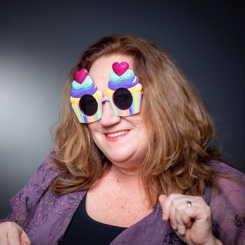 Woman wearing cupcake-shaped glasses with heart-shaped sprinkles. Smiling, posing in front of a dark gray background.