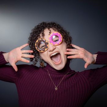 A woman with curly hair wears donut-shaped glasses, yelling with hands raised. Purple turtleneck, gray background.