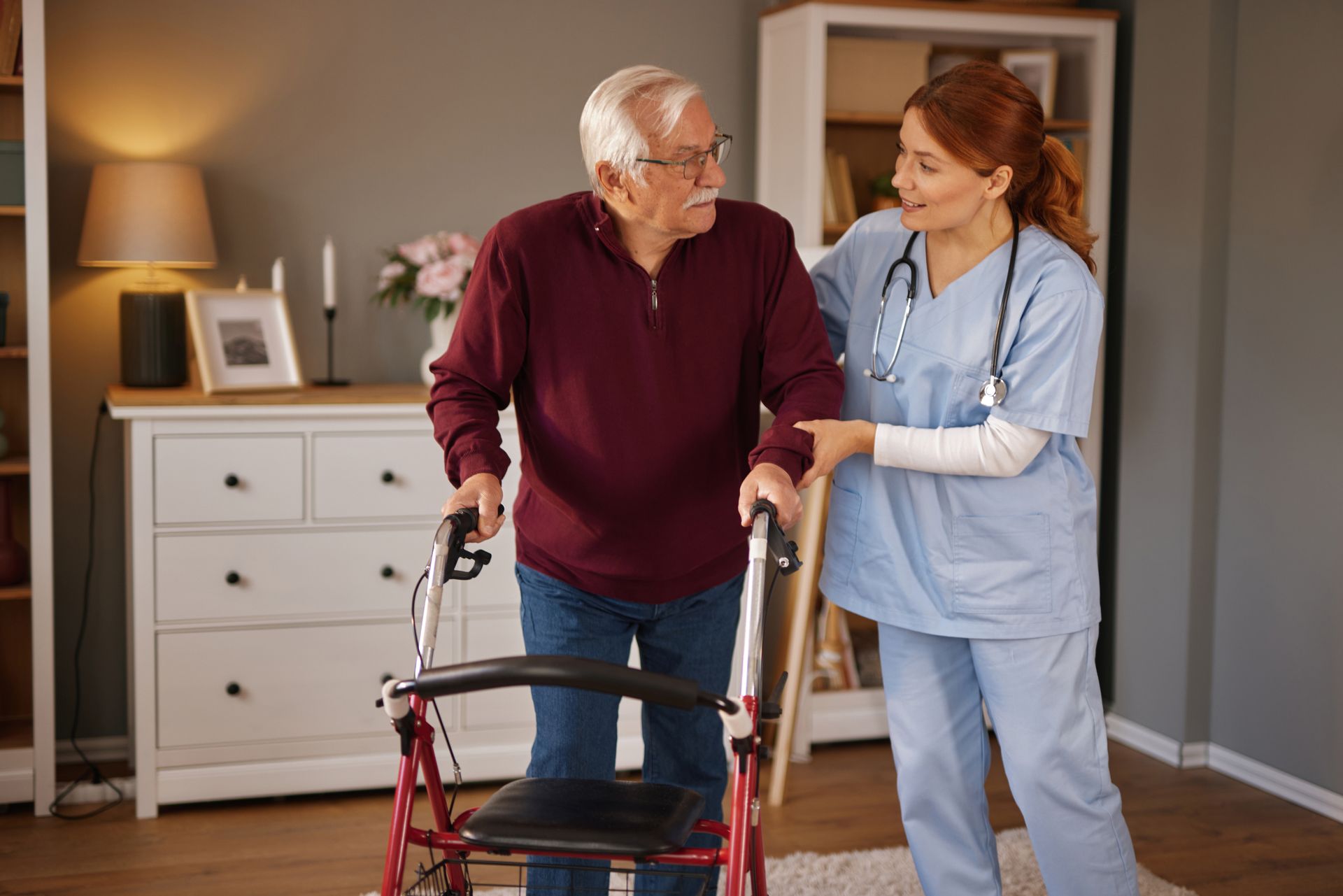A caregiver in blue scrubs assists a person using a red wheeled walker inside a home.