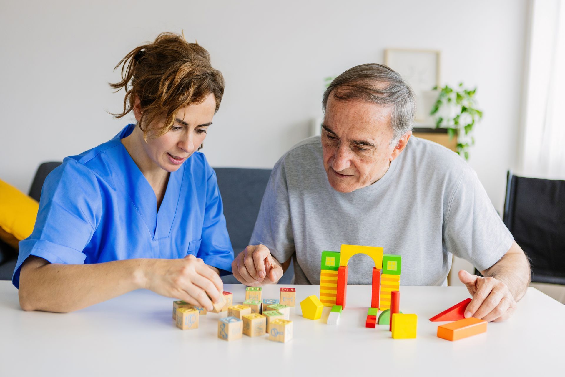 A female caregiver is helping a senior man with wooden shape puzzles for dementia support.
