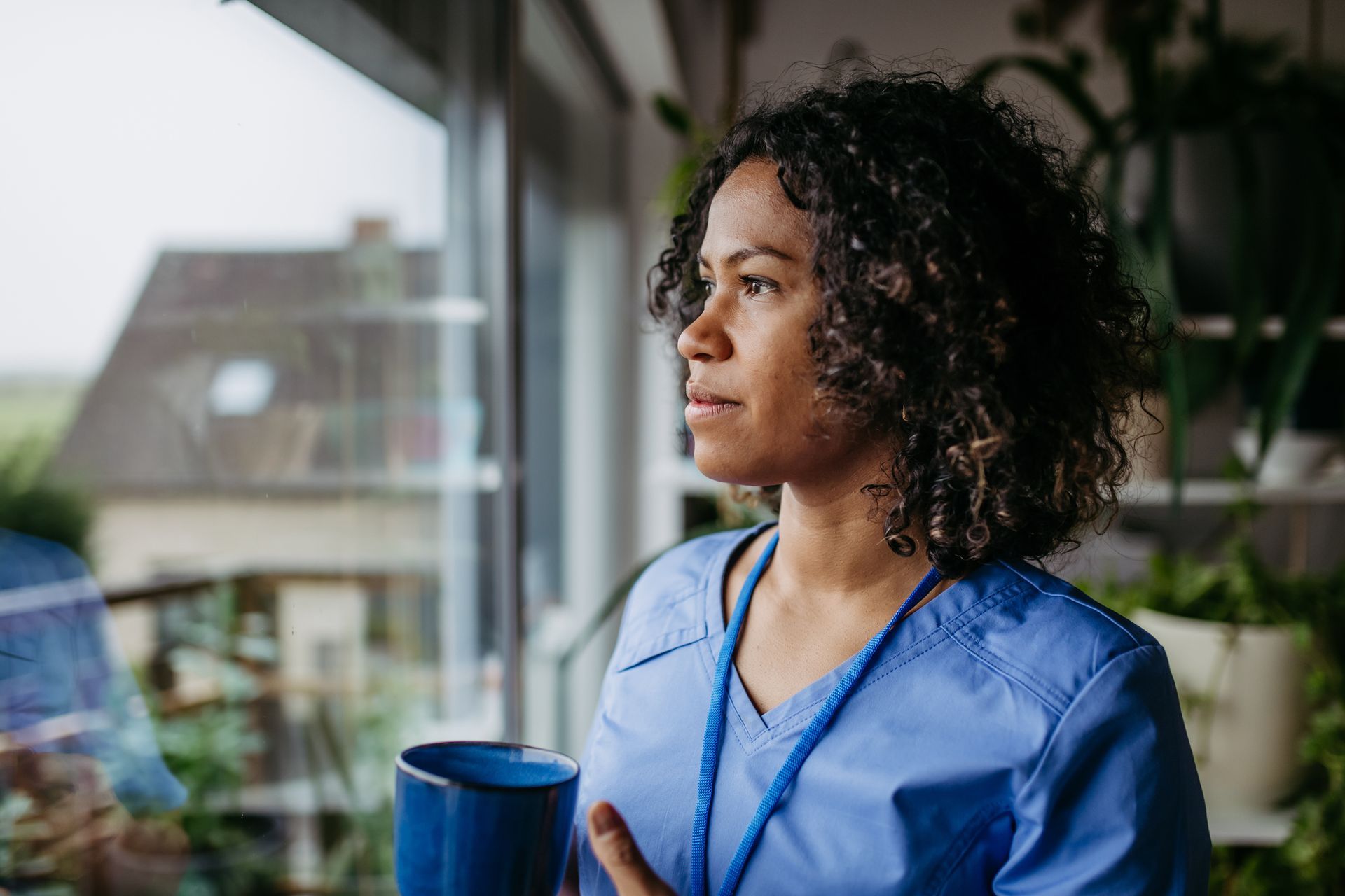 Person in blue scrubs holding a mug and looking out of a window.