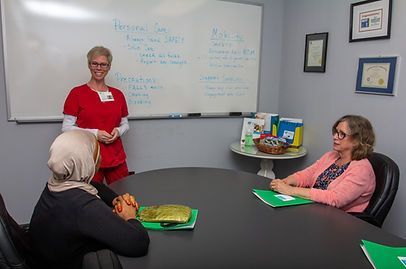 A nurse in red presents to two women seated at a round table. A whiteboard lists notes.