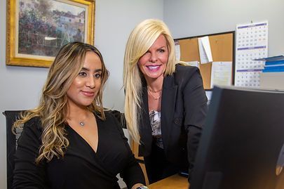 Two women in office setting, looking at computer. One blond, one brunette, both smiling.