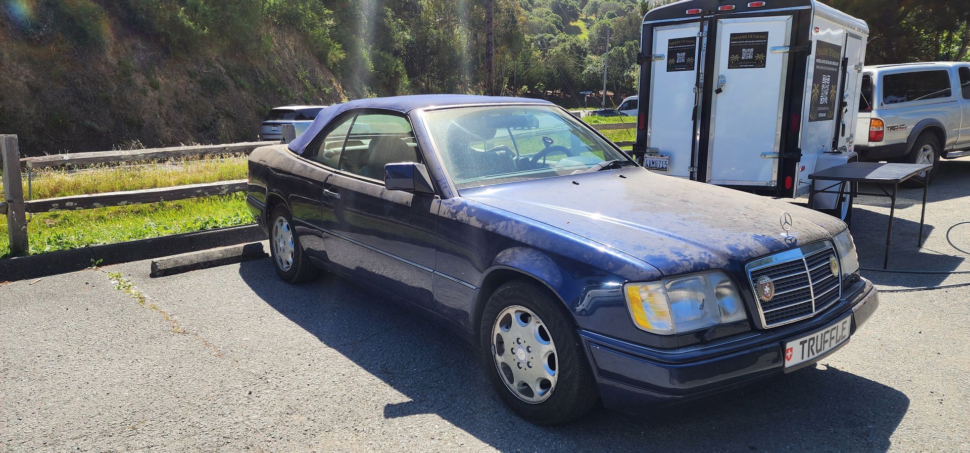 Dark blue sedan parked outdoors beside a white trailer in bright sunlight