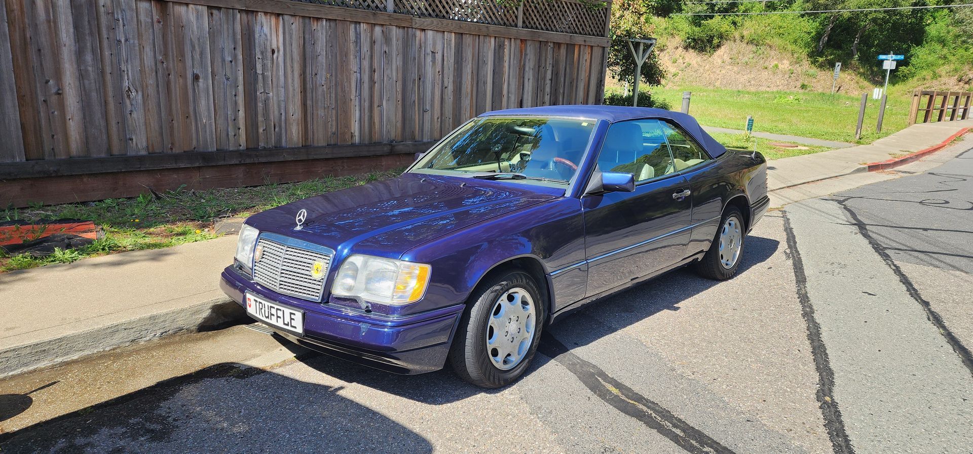 Blue sedan parked on a sunlit street beside a wooden fence and grassy hillside.