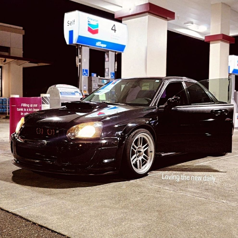Black sports sedan parked at a gas station at night, headlights on.