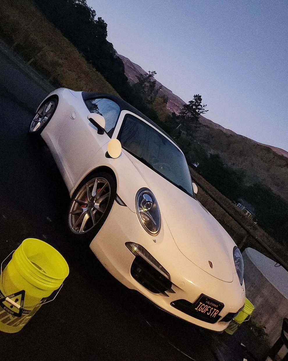 White sports car parked outdoors at dusk beside a yellow bucket on a slanted driveway.