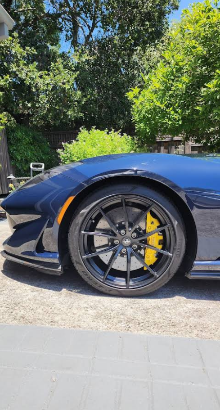 Black sports car close-up with glossy blue paint, black wheel, and yellow brake caliper on a sunny driveway