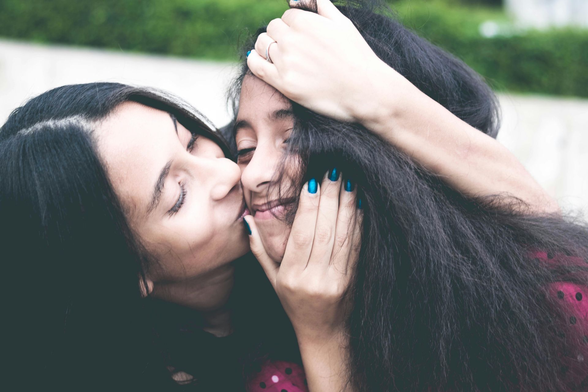 Woman kisses girl on the cheek, both with long dark hair, smiling.