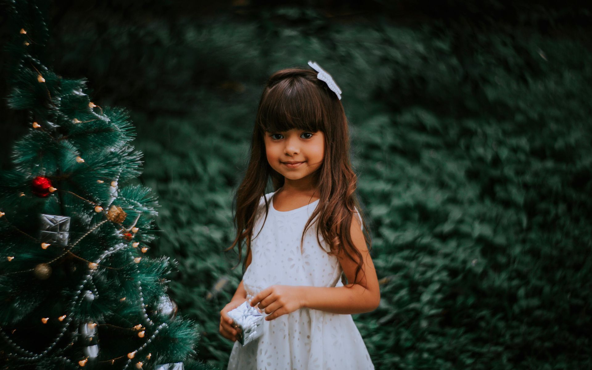 Girl in white dress holding gift, smiling by Christmas tree in outdoor setting.