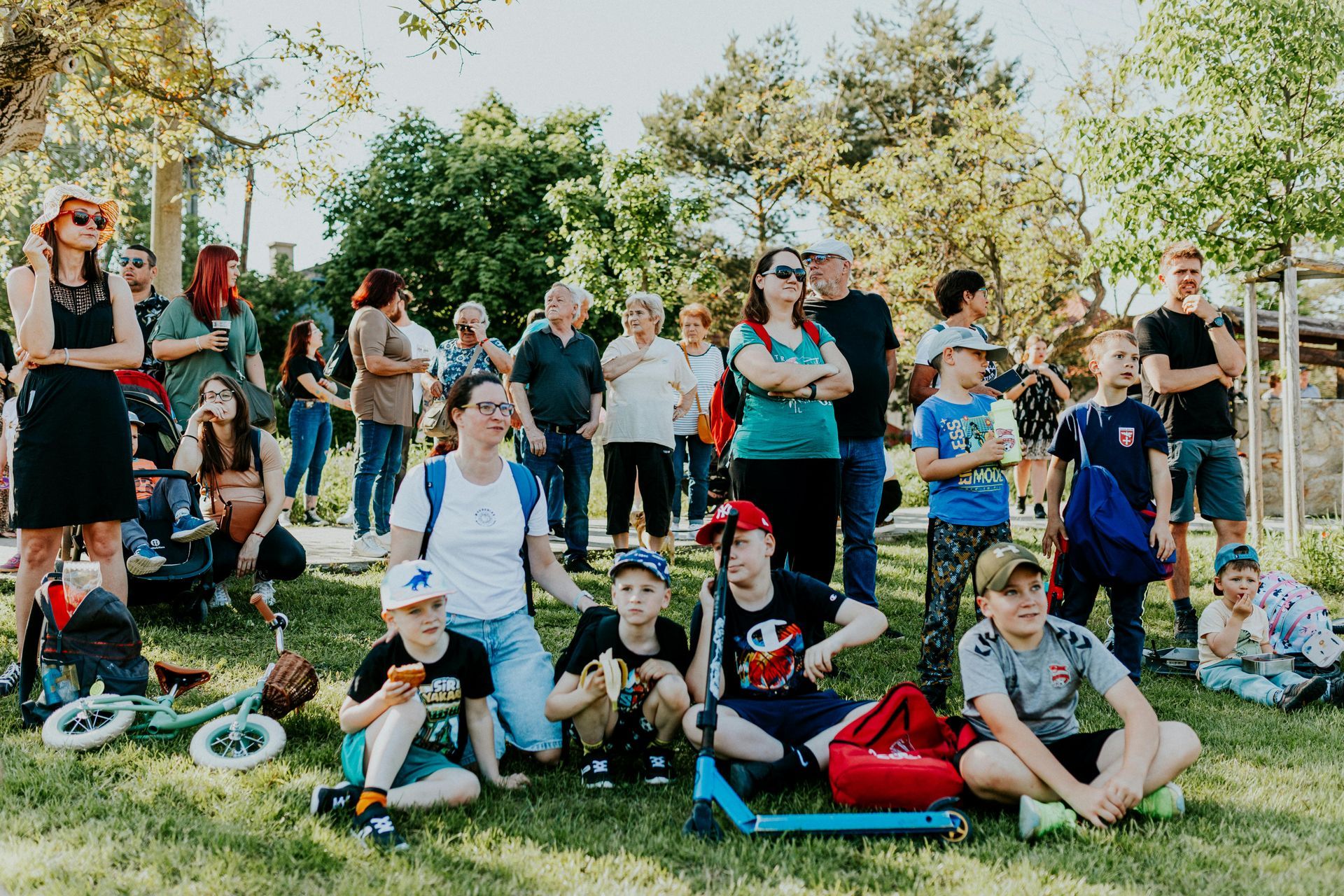 Group of people gathered outdoors in a park, with several seated on the grass and others standing nearby.