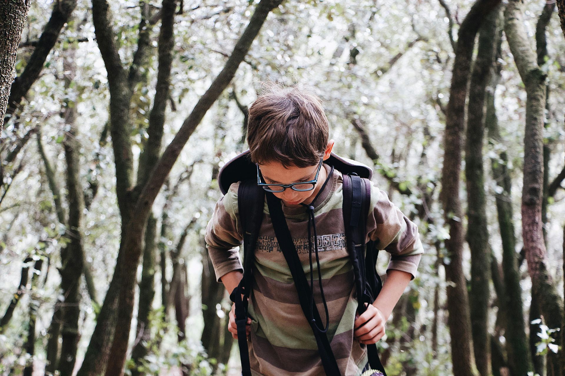 A person wearing glasses and a camo-patterned shirt with a backpack, standing among thin, vertical trees in a forest.