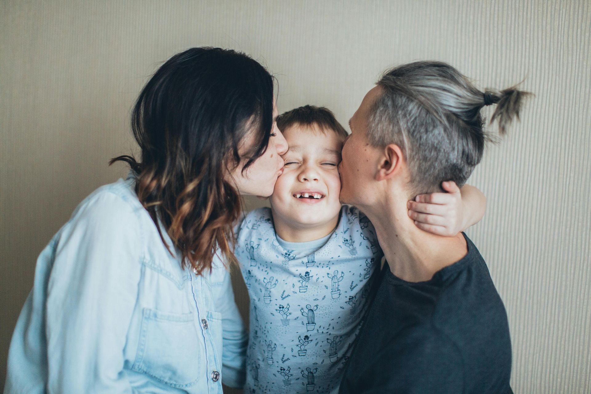 Two adults kissing a child's cheeks; all three are smiling and close together. Neutral background.