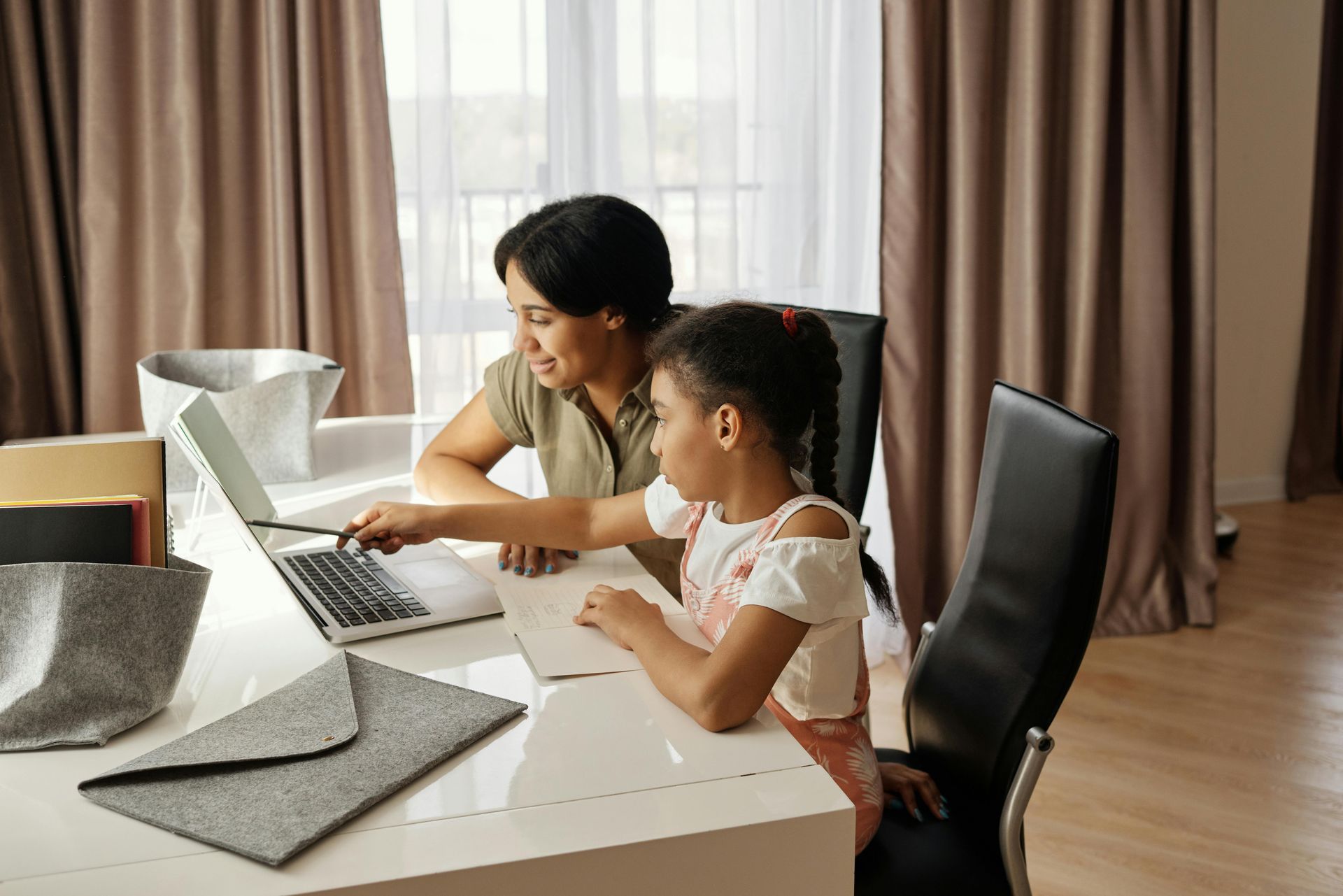 Woman points at laptop screen, helping a child with homework at a white desk in a bright room.