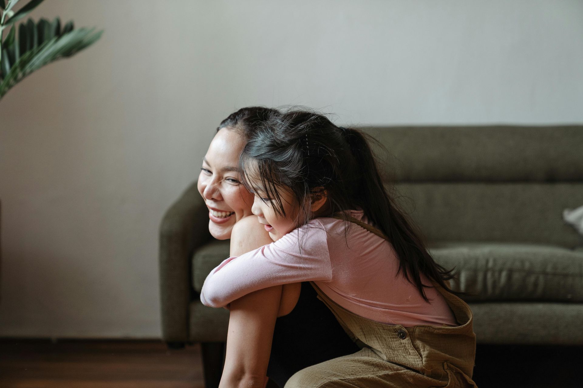Woman smiling, embracing a child indoors near a couch and plant.