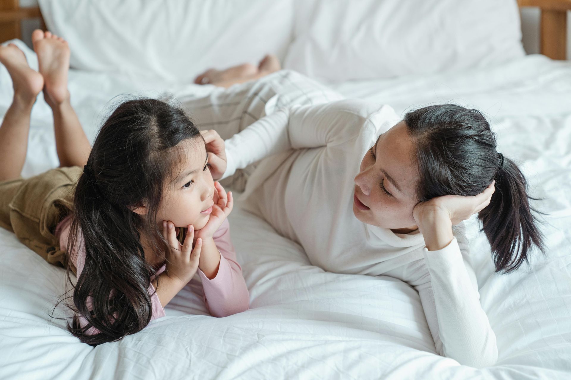 Woman and child lying on a bed, talking. Both are looking at each other, in a light-filled bedroom.