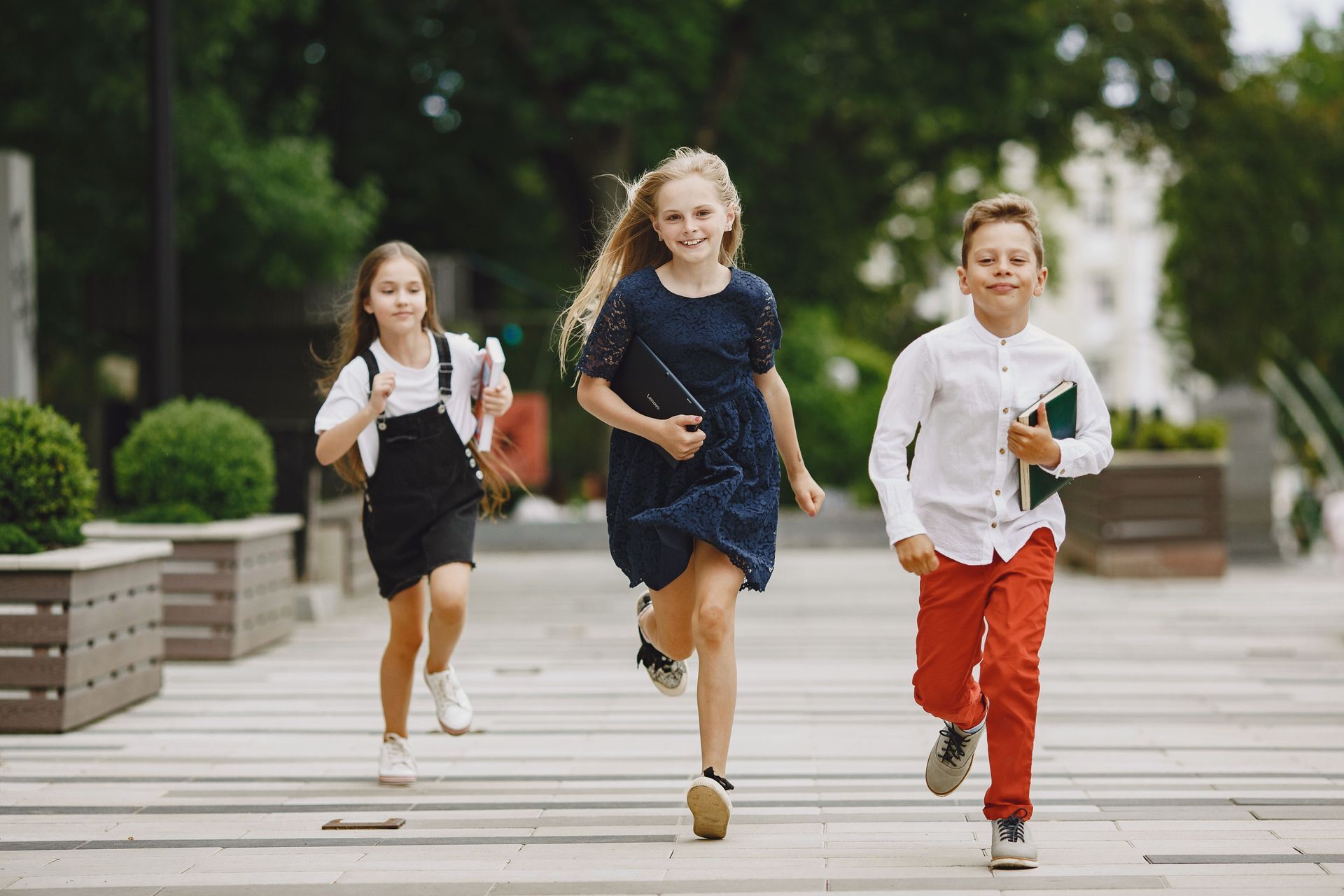 Three children running on a sidewalk in a park, smiling and holding school supplies