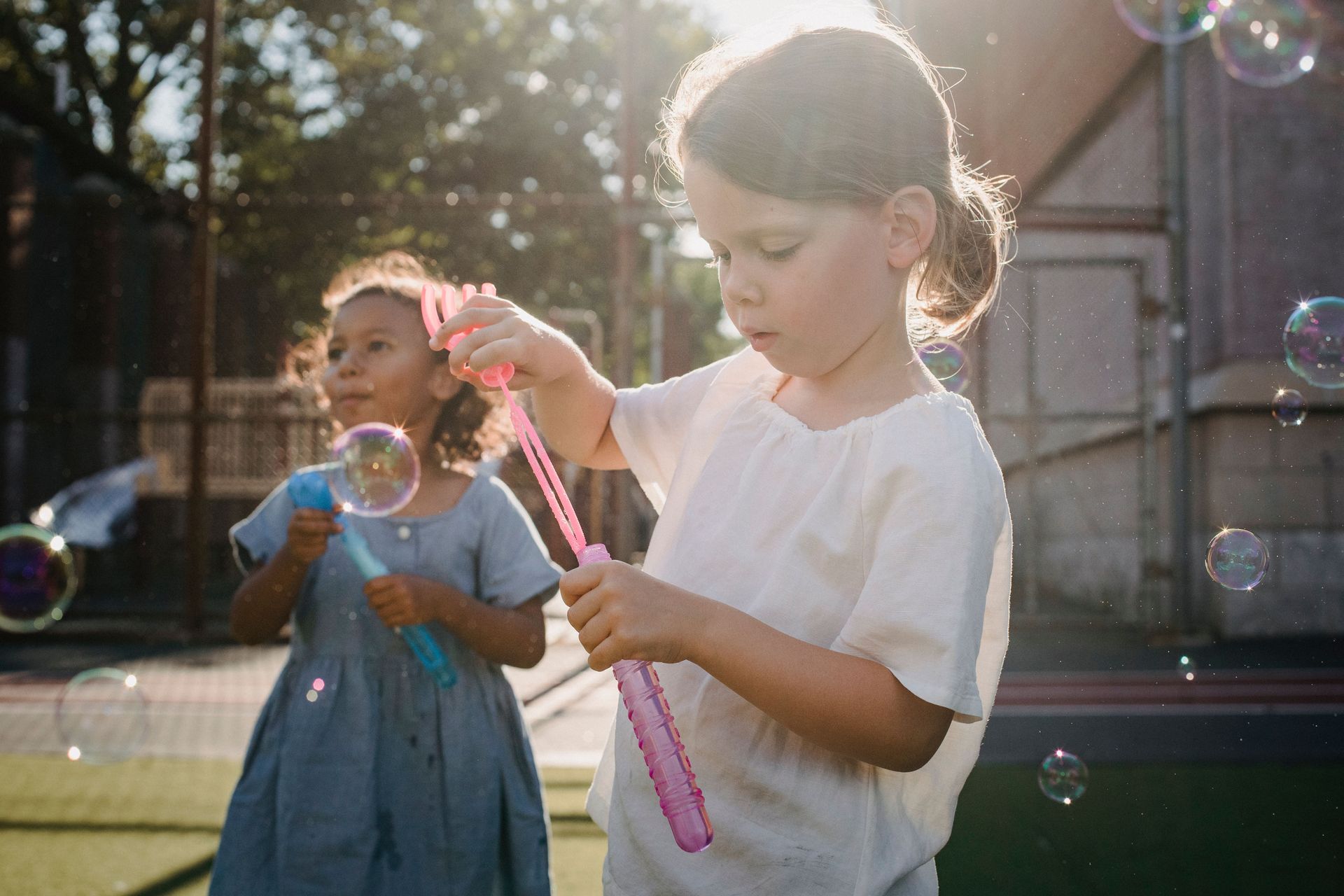 Two children blowing bubbles outdoors in a sunny playground, one holding a pink bubble wand