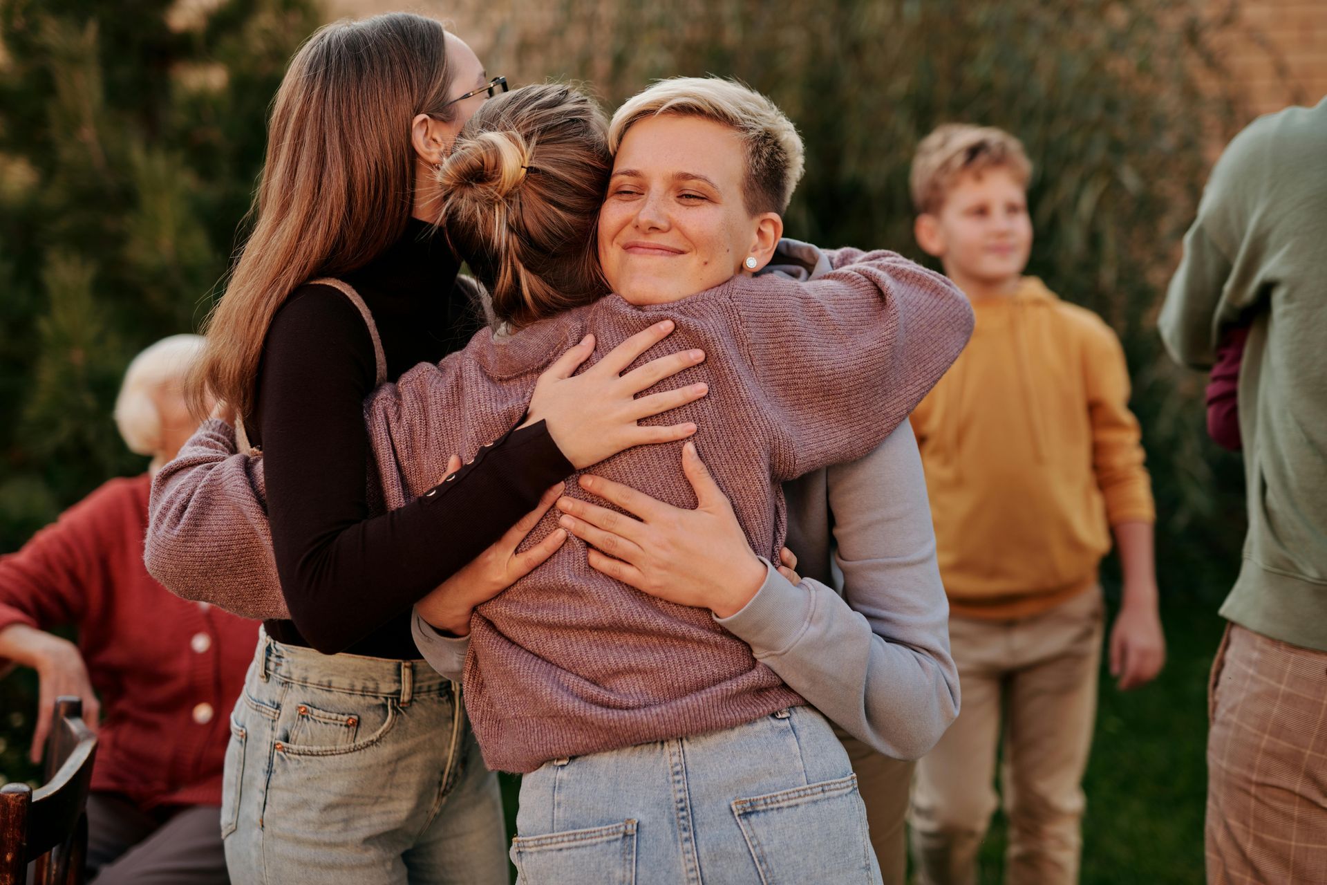 Two people embrace in a backyard. A woman with short hair smiles. Others watch.