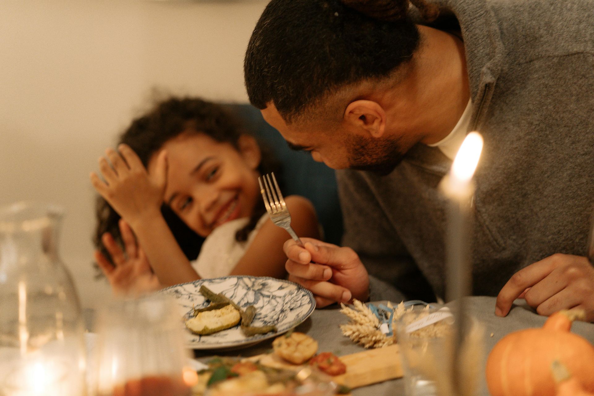A man leans towards a young person smiling at a dining table with food and a lit candle.