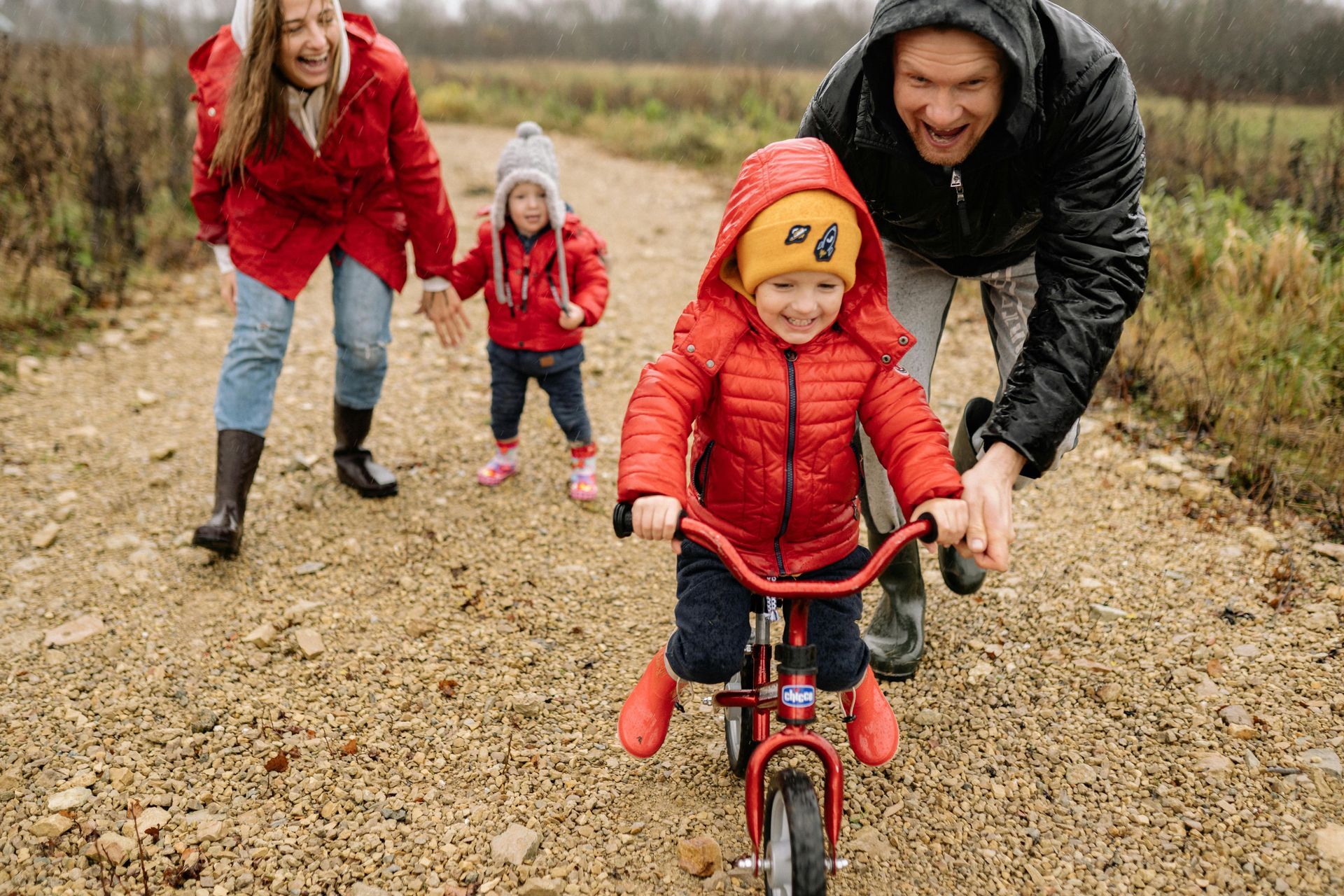 Family helps toddler ride a red bike on a dirt path; mother and father smile as they assist.