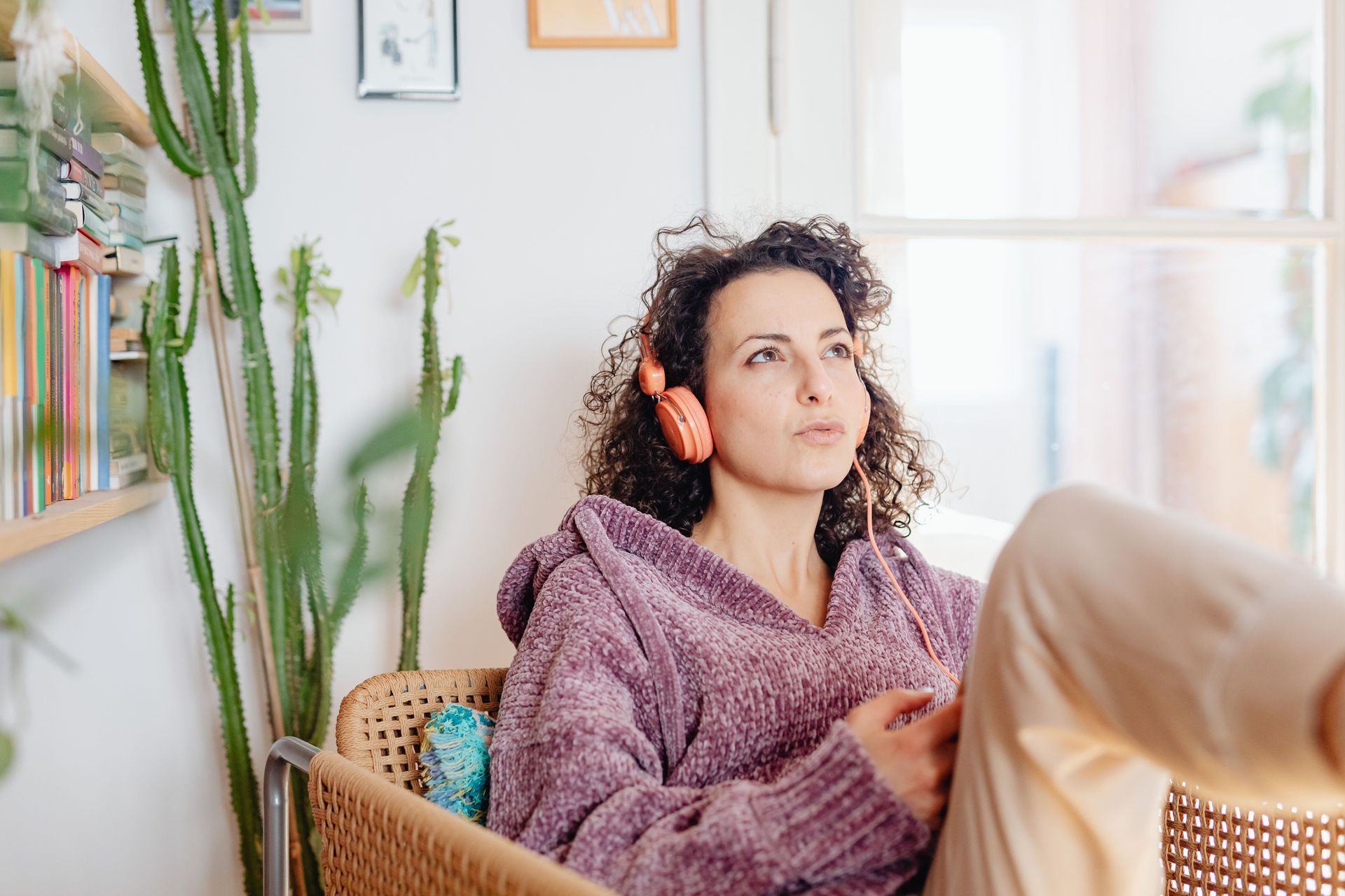 Woman reclining in a wicker chair beside tall cacti in a bright living room, wearing a purple sweater.