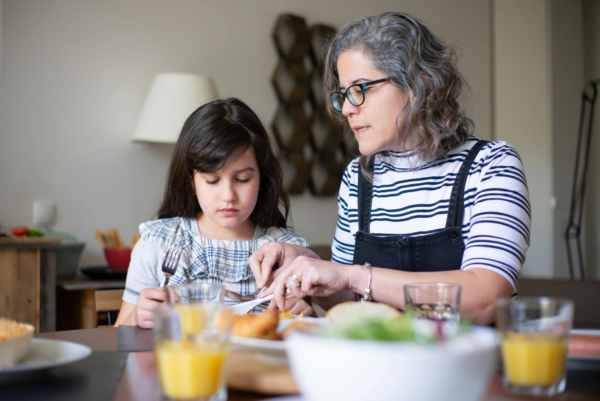 Woman helping child eat at a table. Food and drinks are present.