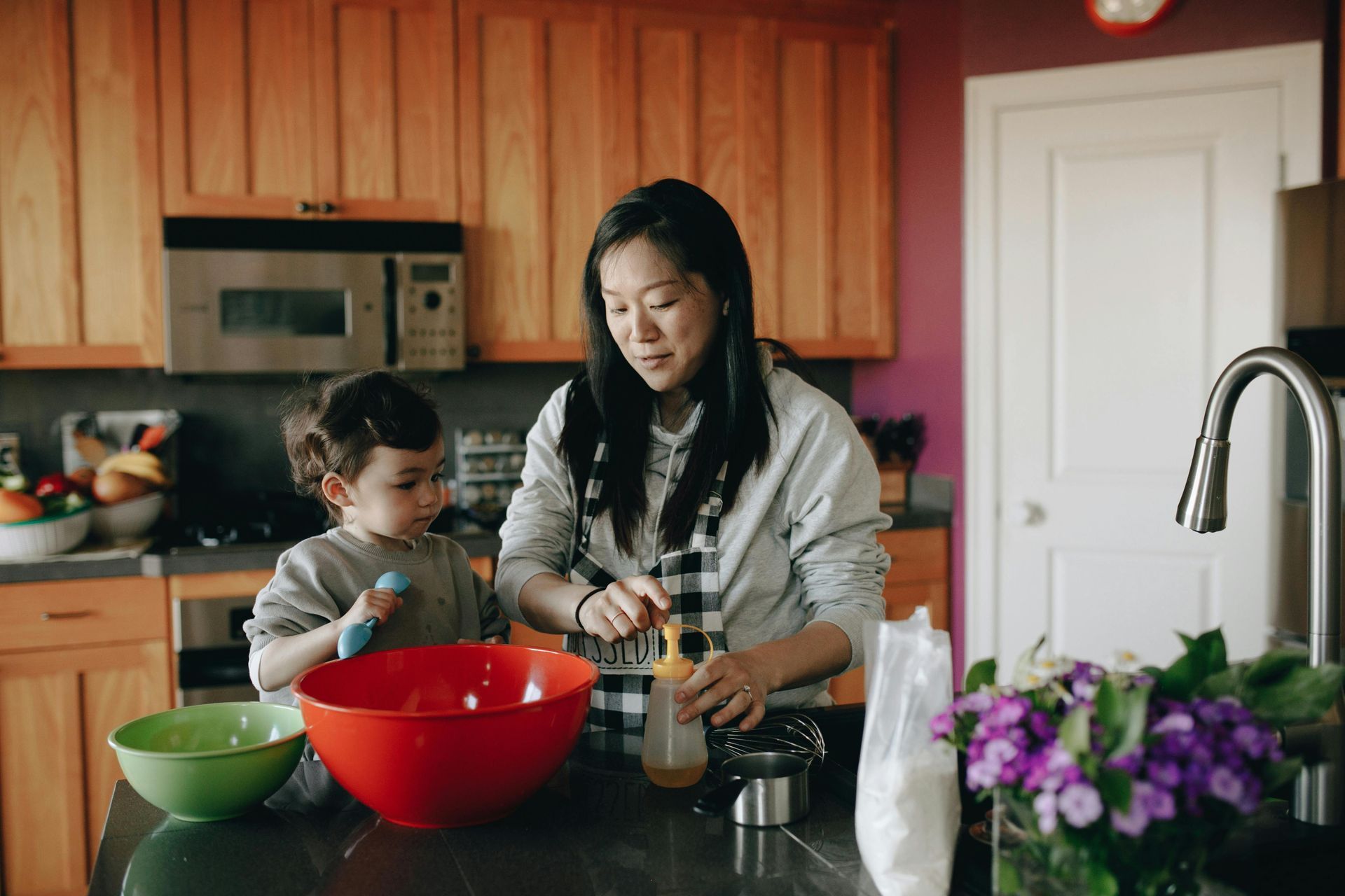Woman and child baking in a kitchen. The woman pours liquid from a bottle, and the child holds a spoon.