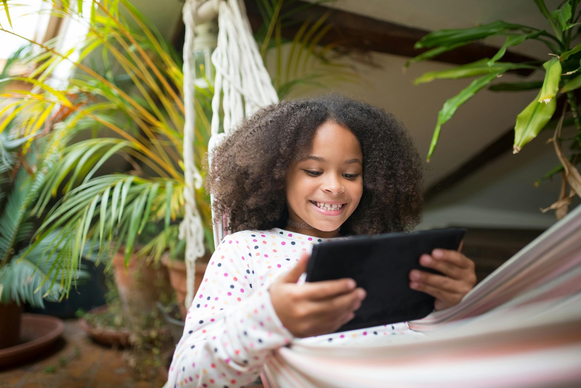Young girl with curly hair smiles, engrossed in a tablet while relaxing in a hammock surrounded by plants.