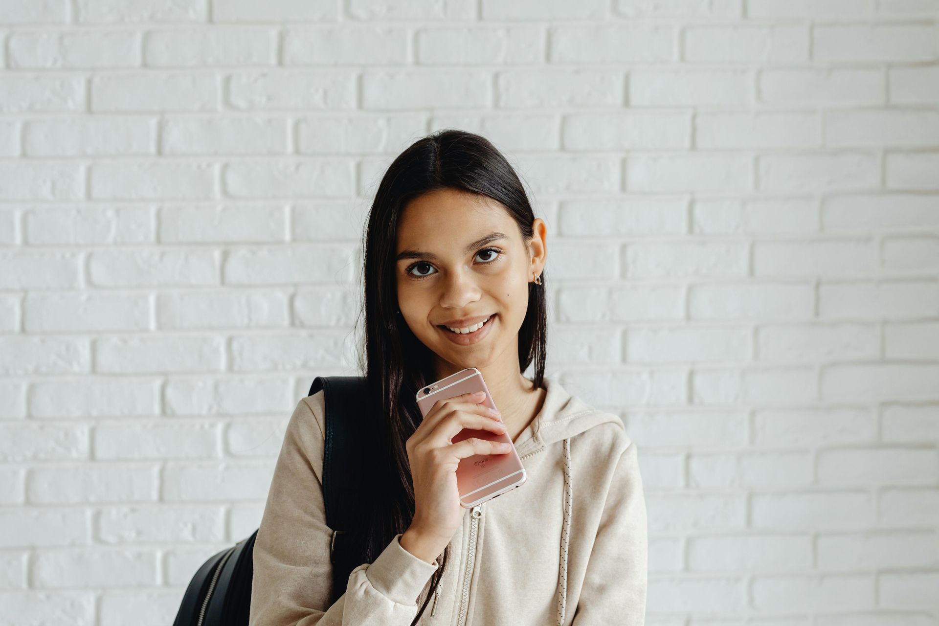 A young girl is sitting in front of a white brick wall holding a cell phone.