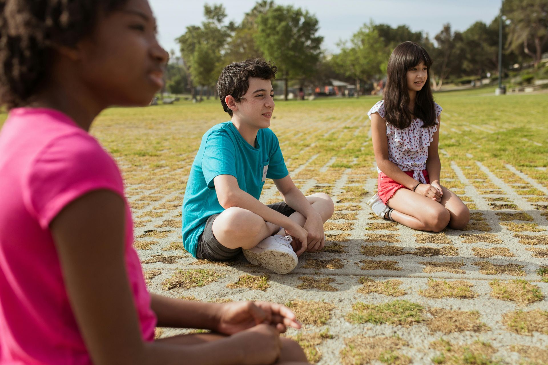 Three children sitting cross-legged on a grassy field outdoors, surrounded by park landscaping