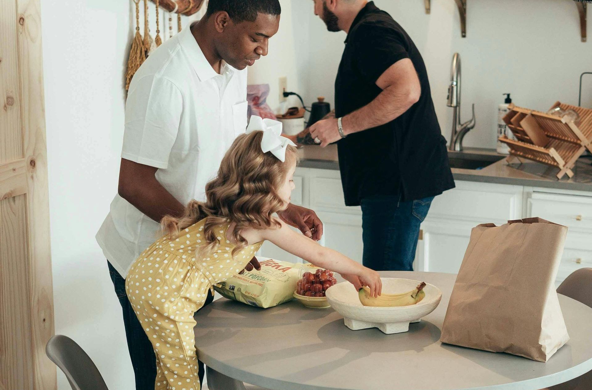 Two men and a little girl are standing around a table in a kitchen.