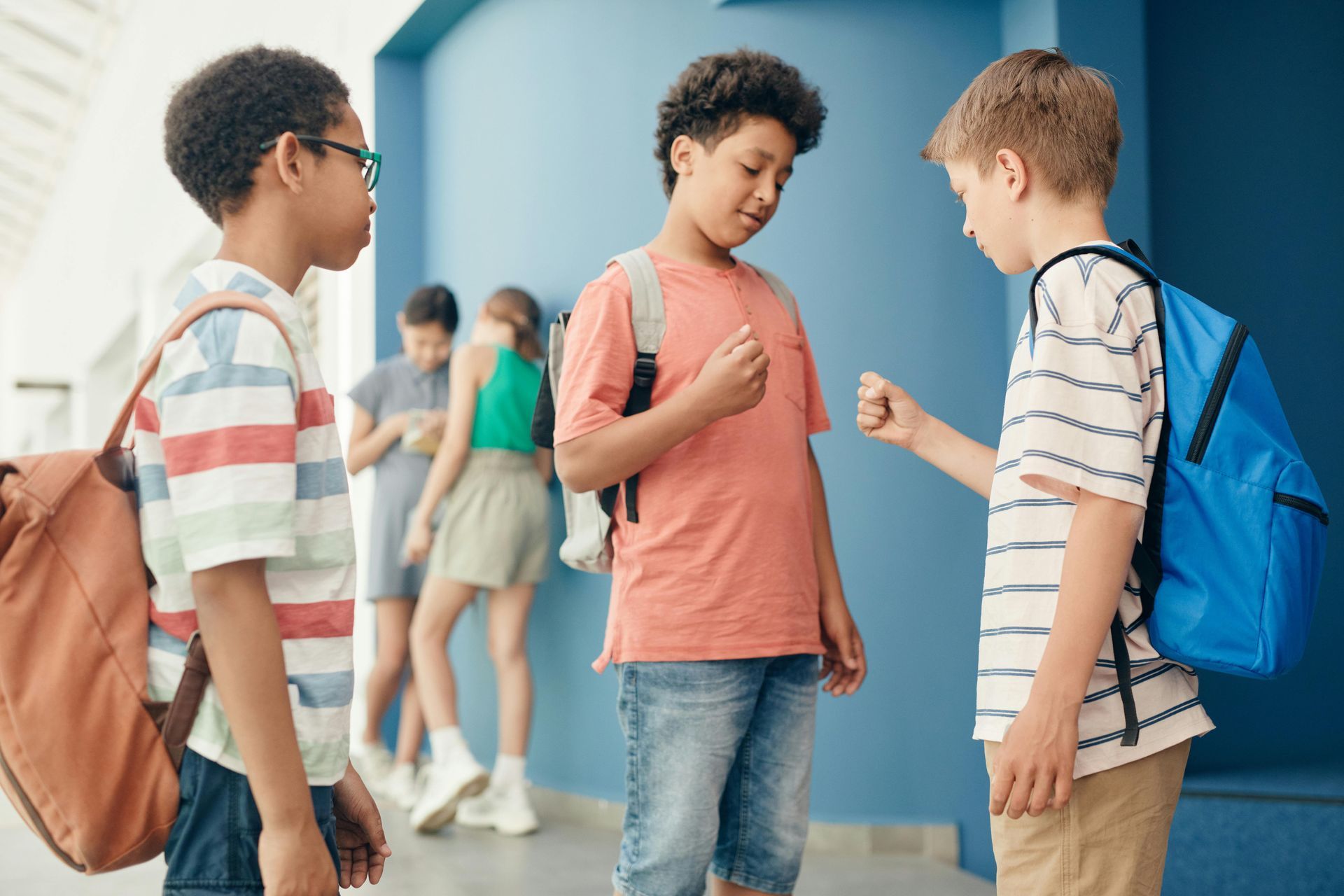 Three students talking in a school hallway, with backpacks and lockers in the background.
