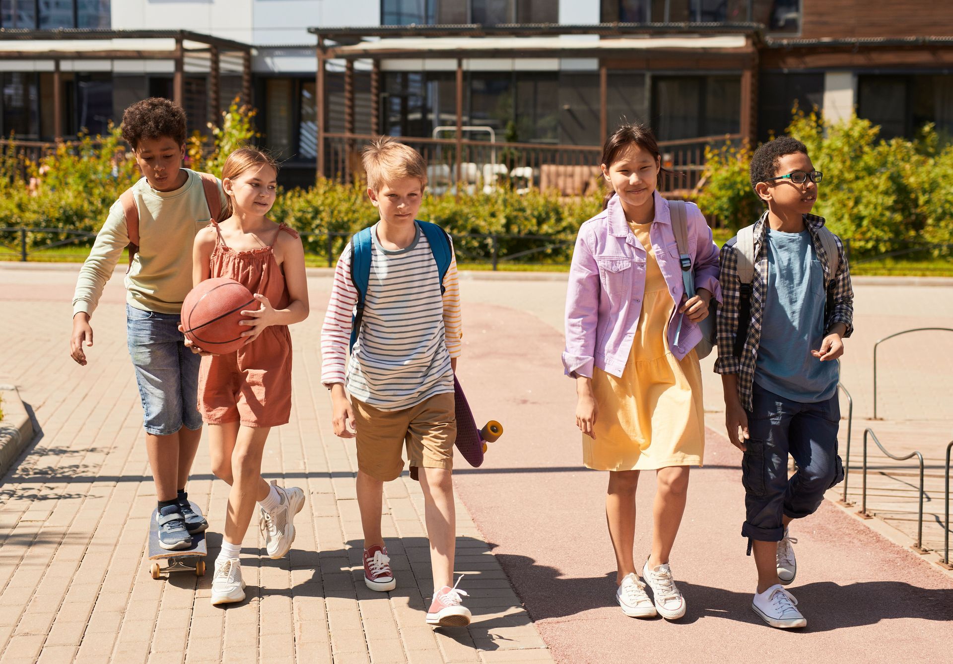 Five children walking together outside a school building on a sunny day