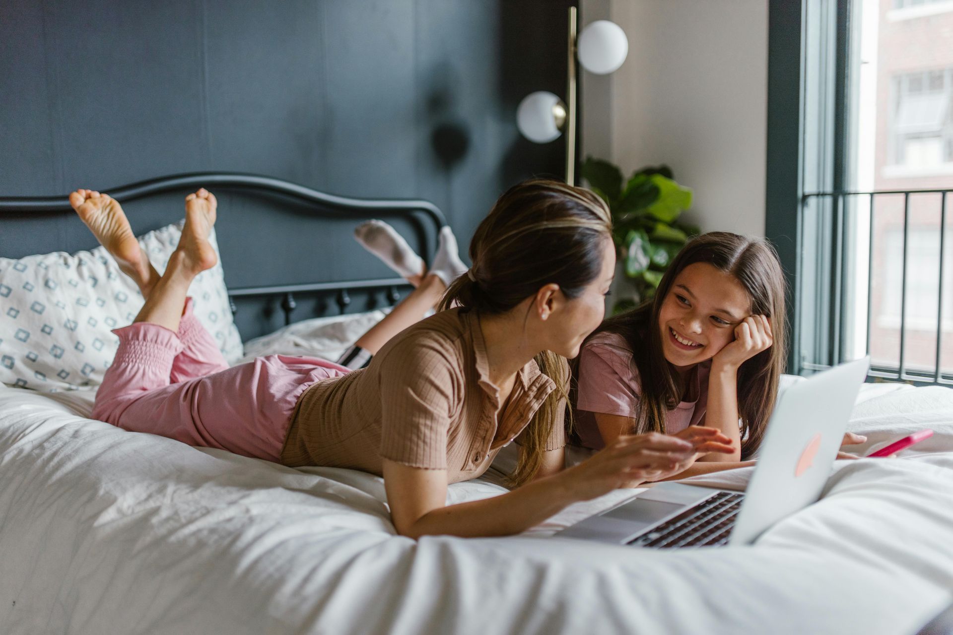 Two people lying on a bed, smiling and using a laptop in a bright bedroom