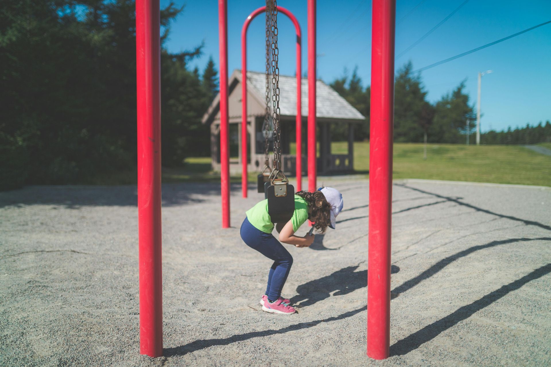 A child in a bright green shirt stands stooped under a red swing set in a playground with a small wooden shelter behind.