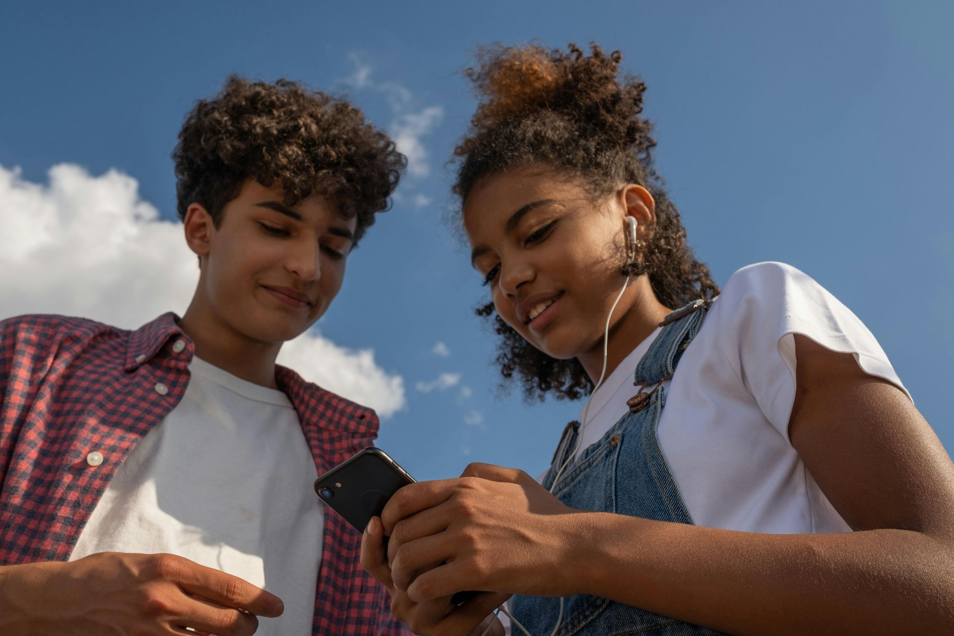 Two teens looking at a smartphone outdoors under a blue sky