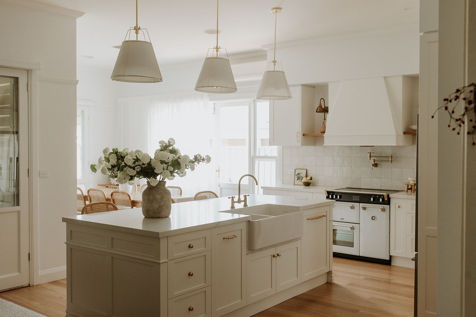 Light-filled white and modern country kitchen with spacious stone island bench top including a double farm house sink, painted shiver profile drawer fronts, pull out bin and brass tapware and knobs and handles. Cooktop wall has ornate canopy with mantle concealing the rangehood, tile splashback and pot taps.