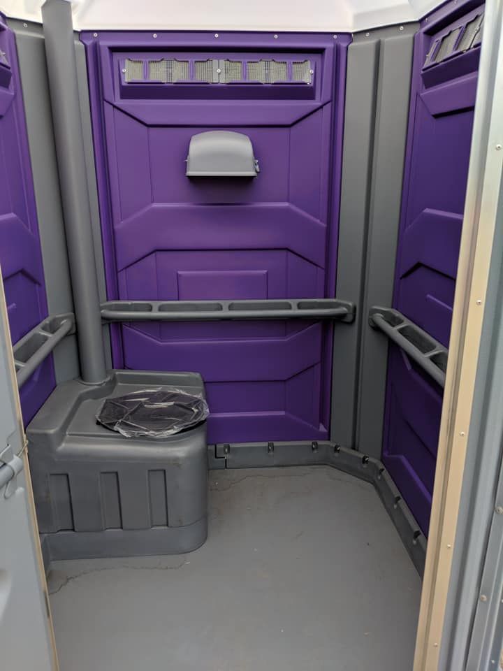 A purple and gray portable toilet with a trash bag on the floor.