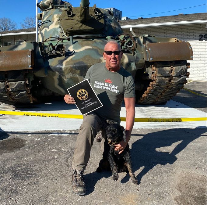 A man is sitting next to a dog and holding a certificate in front of a tank