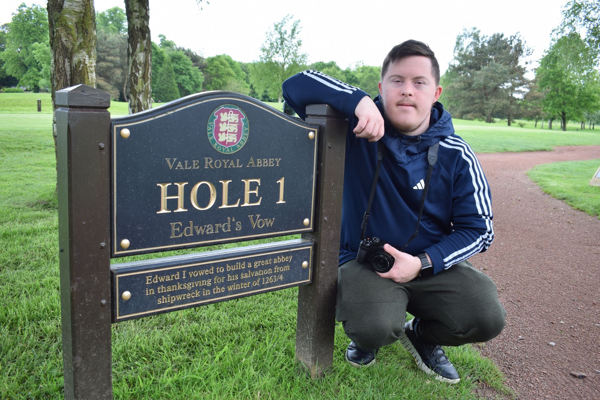 Young adult with Down syndrome crouching next to sign saying Hole 1 of Vale Royal Golf Club