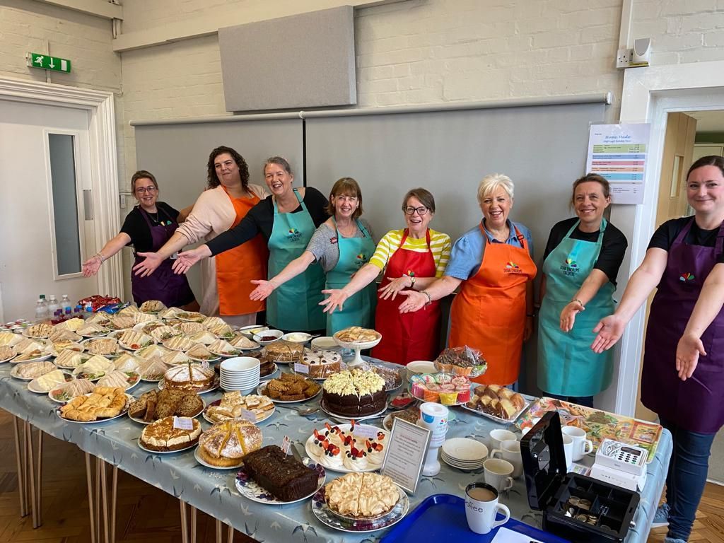 A line of lady volunteers with outstretched arms pointing to a afternoon tea feast for Down Syndrome