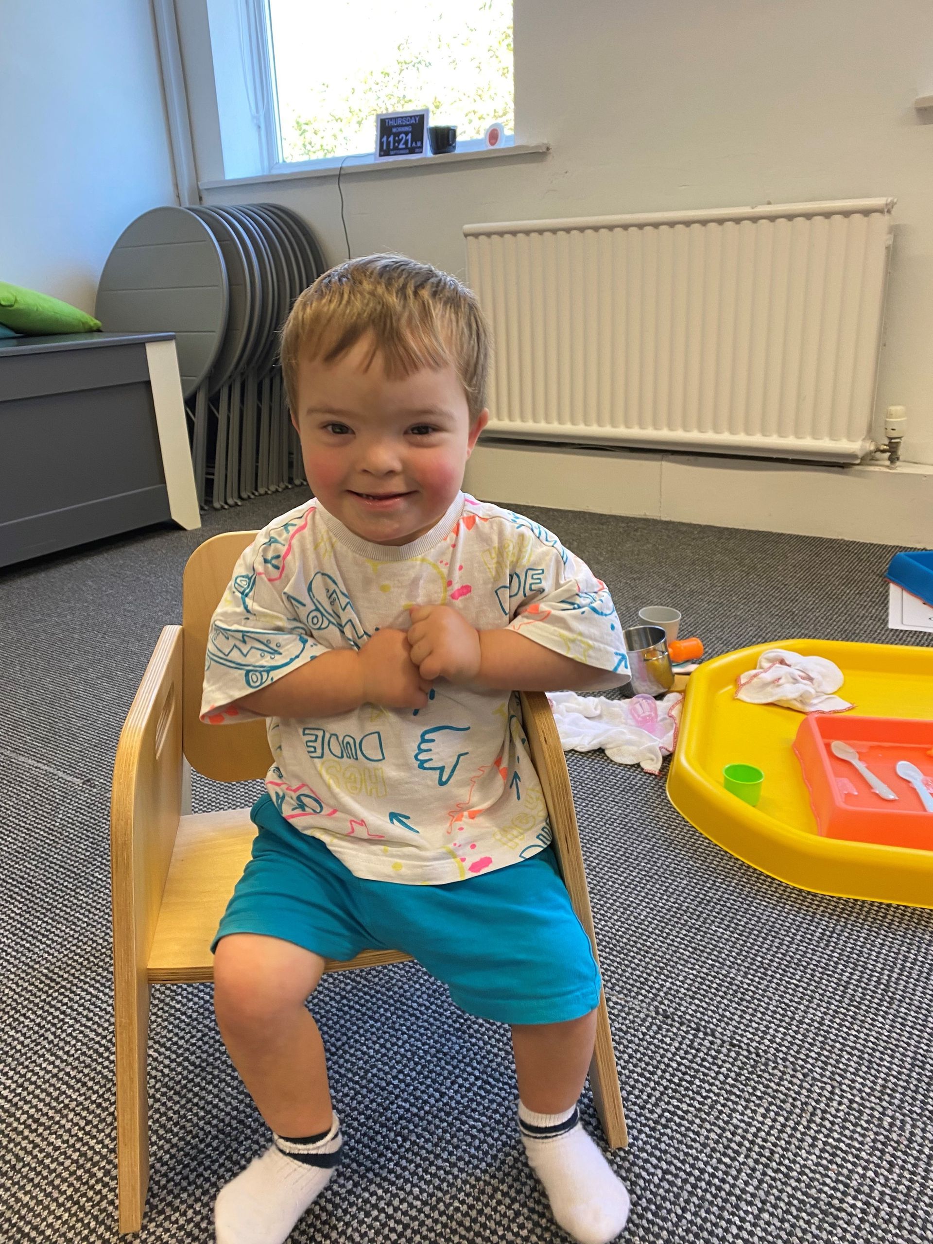 A toddler with Down syndrome seating in a small wooden chair at Down Syndrome Cheshire.