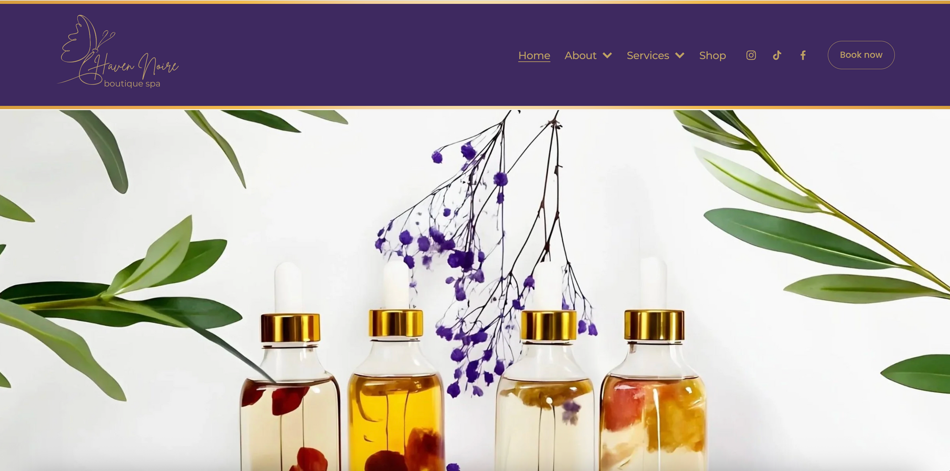 Four glass bottles with gold lids, filled with oils and botanicals, with purple and green foliage.